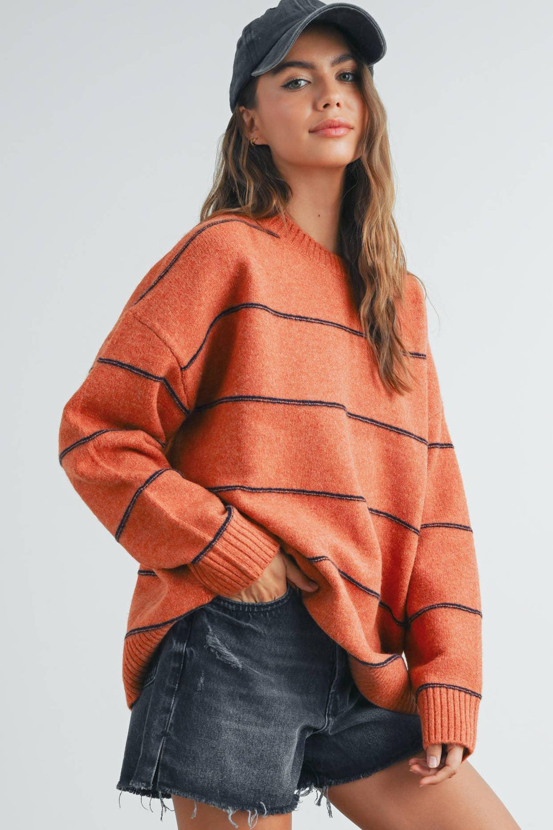 Woman in orange striped sweater, black denim shorts, and baseball cap