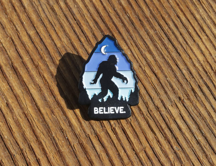 Believe Bigfoot enamel pin with moon and forest design on wooden surface