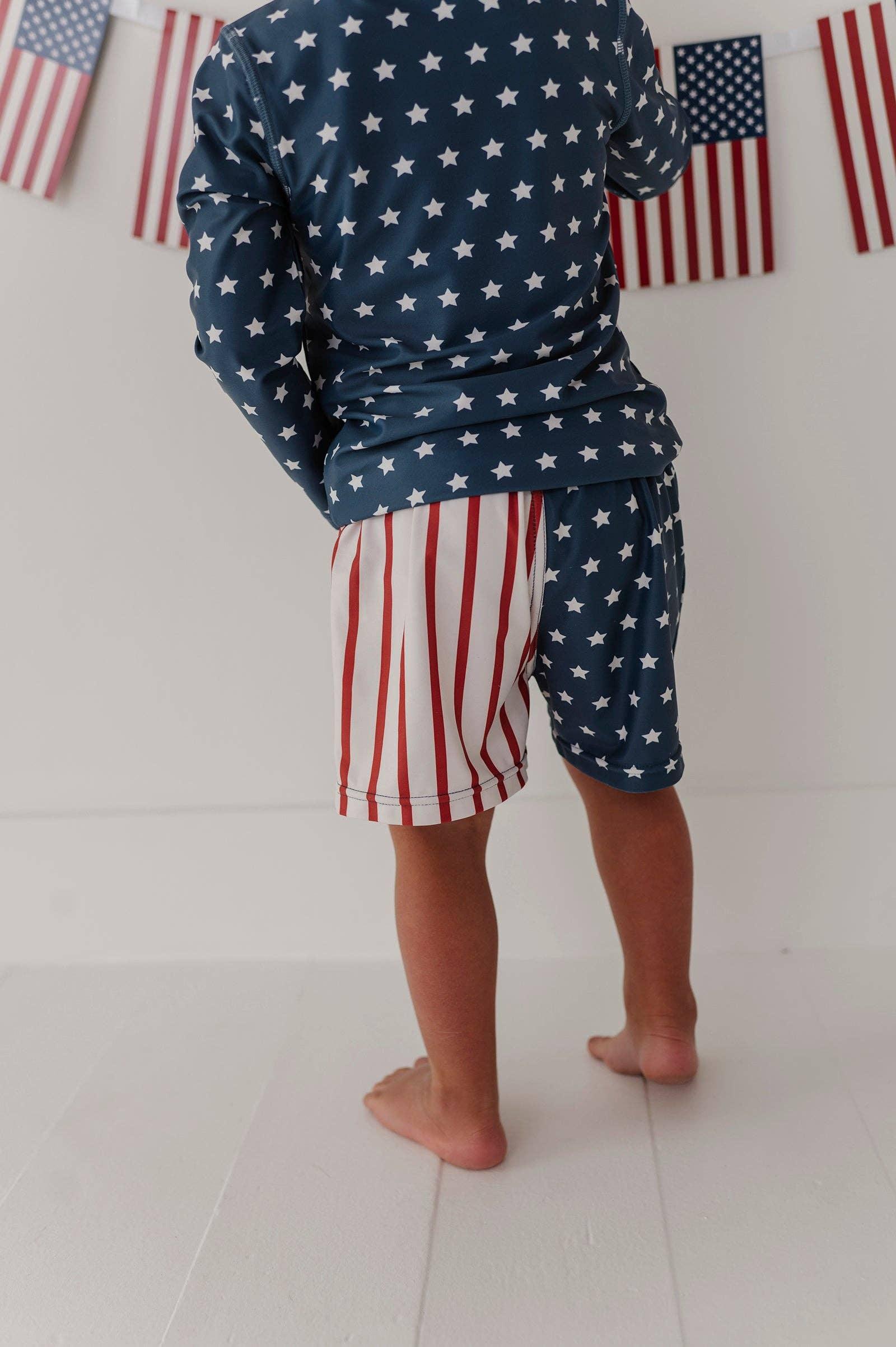 Child wearing stars and stripes swim set with American flag backdrop, perfect for beach days and coastal adventures.