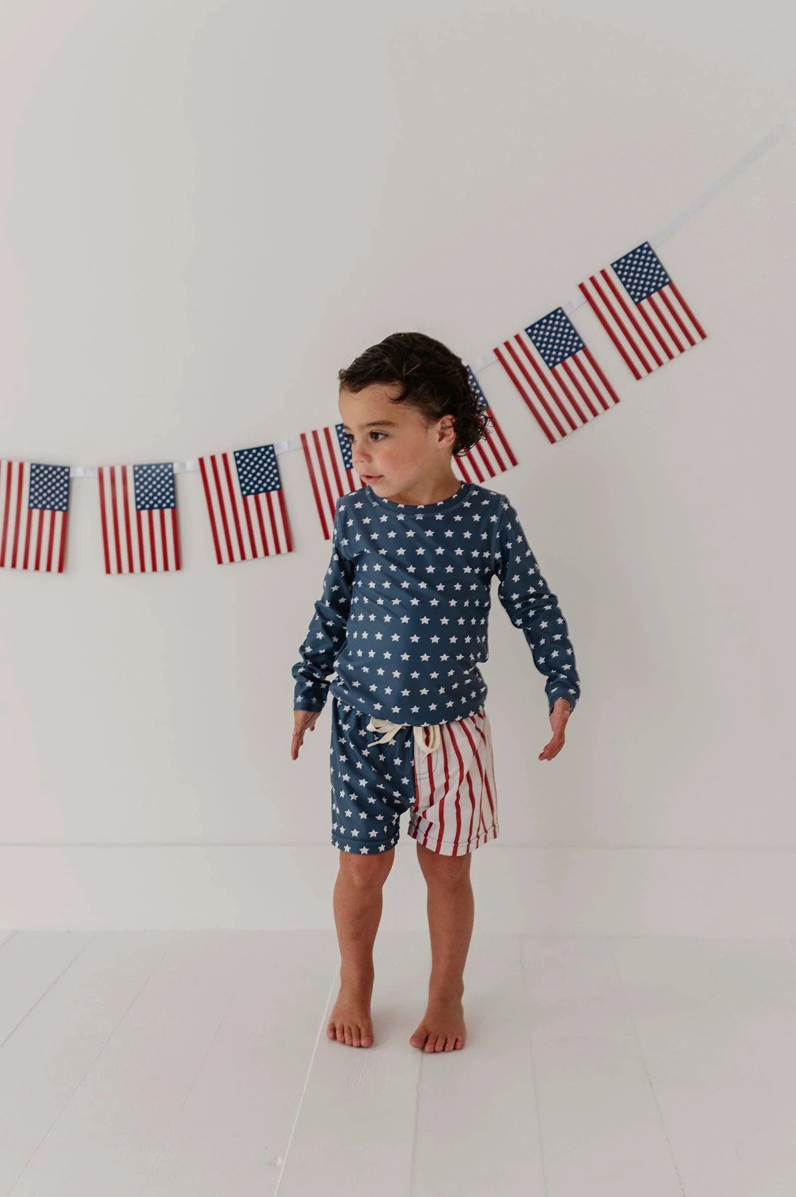Toddler in stars and stripes swim set with American flag banner, perfect for beach days and coastal adventures.