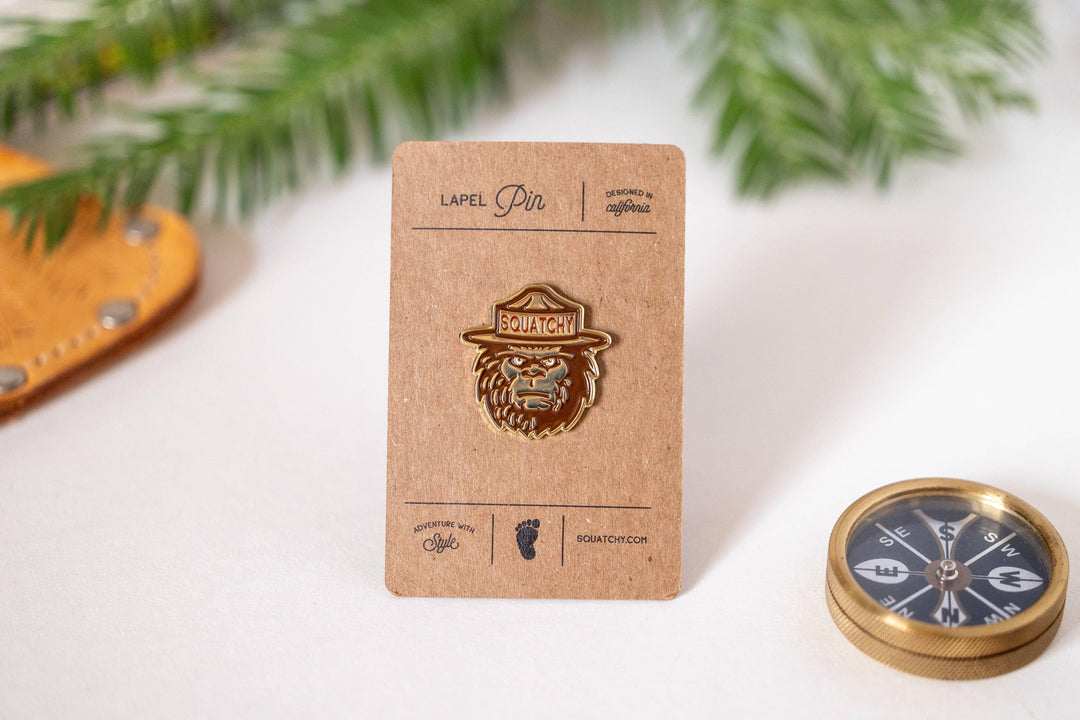 Squatchy lapel pin with hat design, styled with compass and greenery for adventure-themed decor