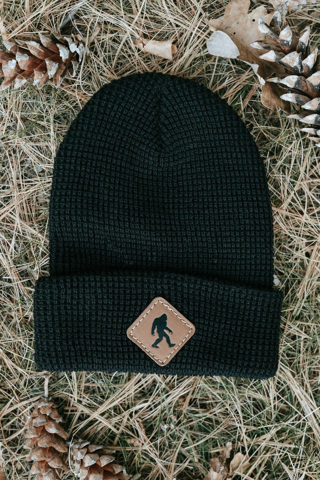 Black waffle knit beanie with Bigfoot patch, styled on pine needles and cones for PNW outdoor adventures