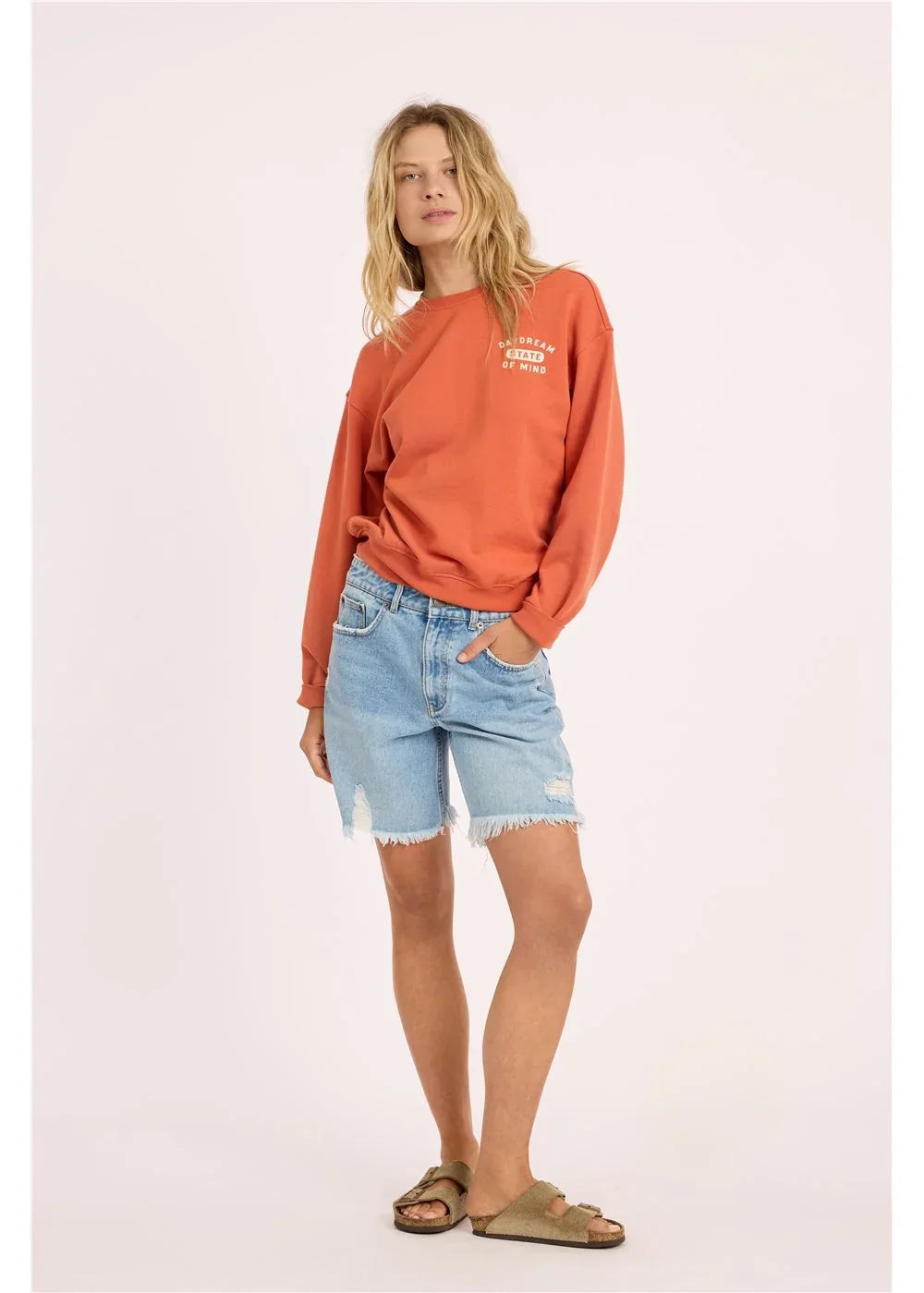Woman in orange graphic sweatshirt, distressed denim shorts, and sandals for coastal PNW style