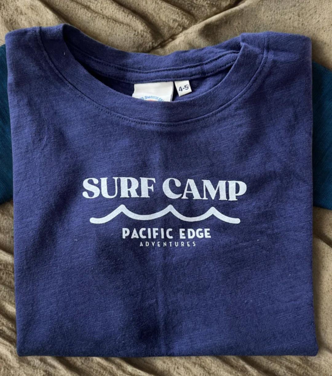 Navy blue kids' Surf Camp t-shirt with Pacific Edge Adventures print, folded on cozy fabric