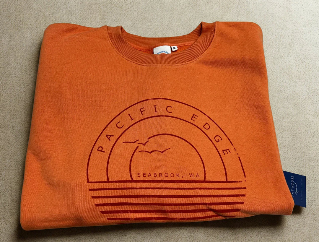 Orange crewneck sweatshirt with 'Pacific Edge Seabrook, WA' graphic, coastal design for PNW outdoor lifestyle