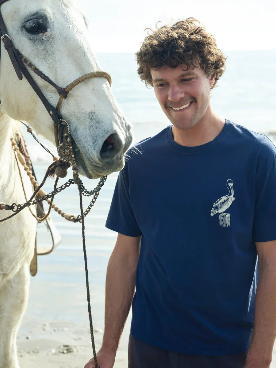 Man in navy pelican tee smiling beside white horse on beach, coastal lifestyle apparel