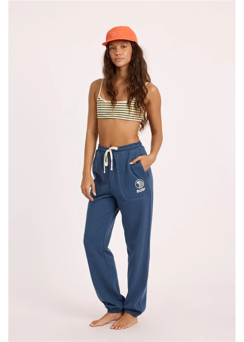 Woman in blue surf joggers, striped crop top, and orange cap — coastal lifestyle gear