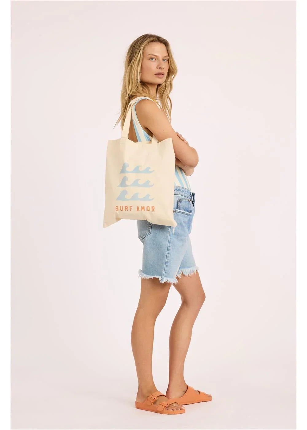Woman in denim shorts carrying Surf Amor tote bag with wave design, coastal style ready for beach days.