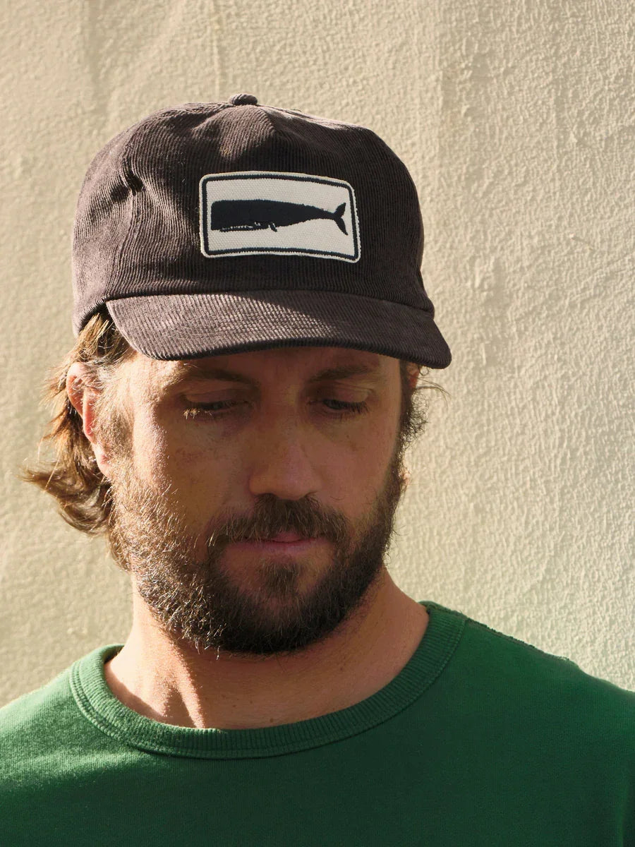 Man wearing dark corduroy cap with whale patch, green shirt, outdoor casual style