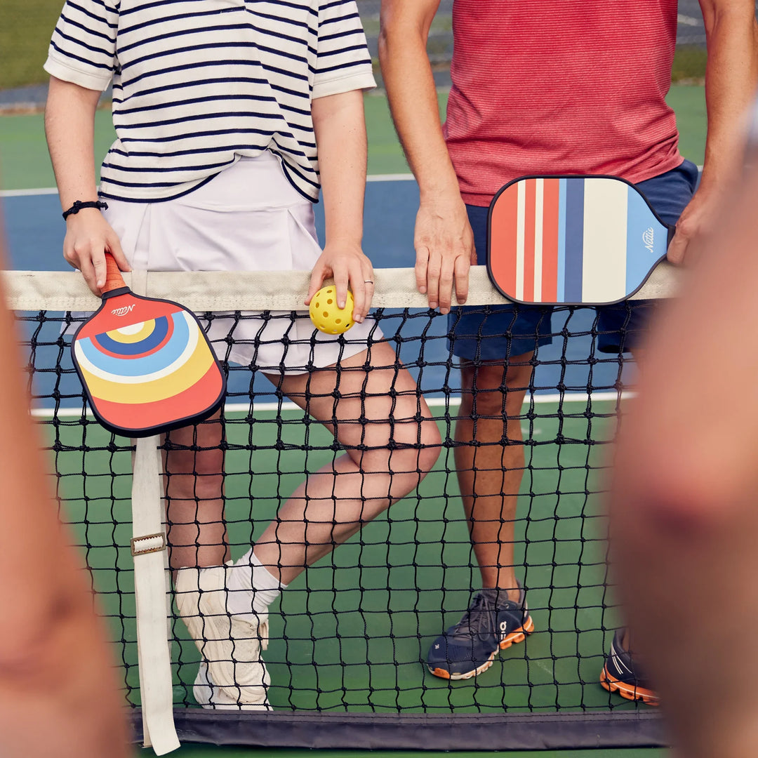 Couple holding colorful pickleball paddles and ball at net on outdoor court, ready for game