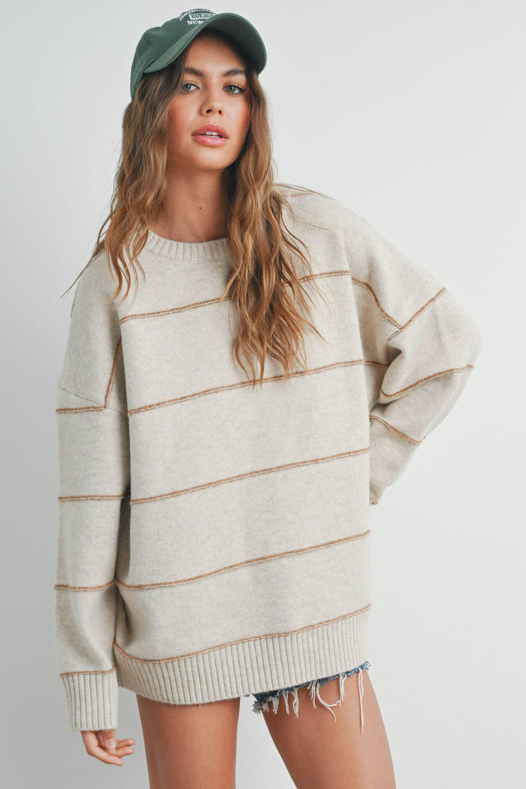 Oversized cream sweater with brown stripes, paired with denim shorts and a green cap
