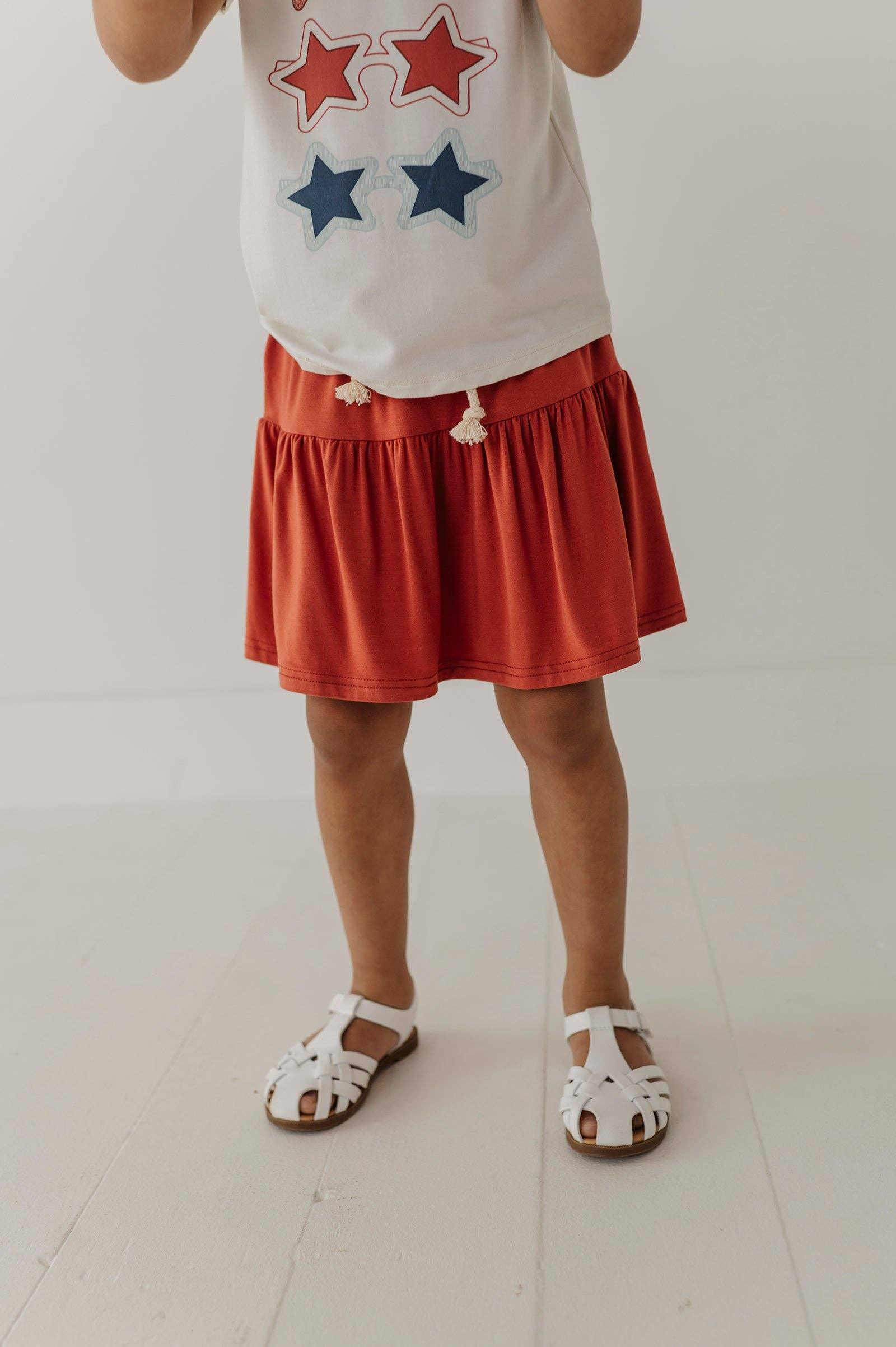 Child in star-print tee, rust tiered skirt with tassels, and white sandals for coastal summer adventures