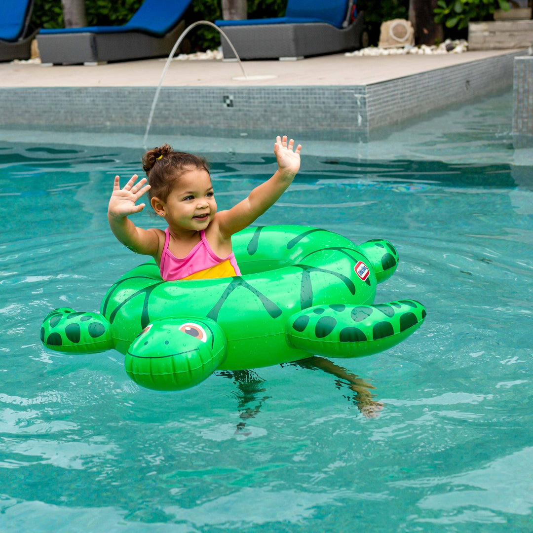 Toddler on green turtle float in pool, waving happily during summer swim fun.