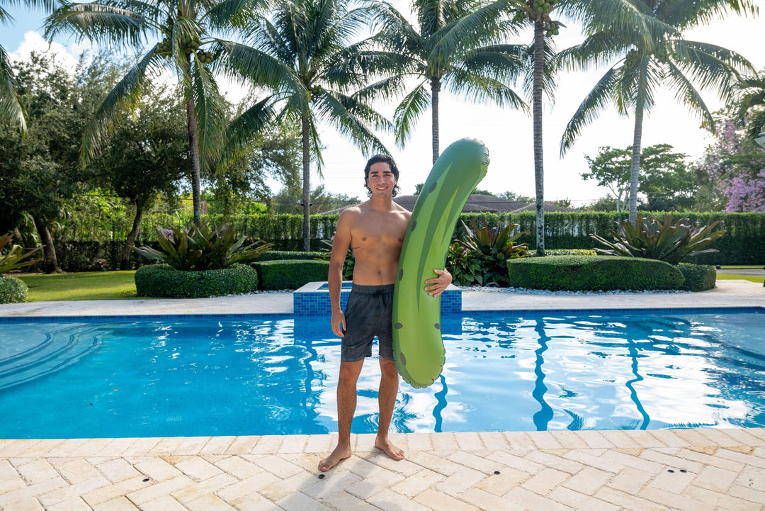 Man holding green pickle-shaped pool float by backyard pool with palm trees, perfect for summer fun and beach days.