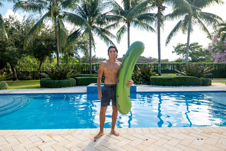Man holding green pickle-shaped pool float by backyard pool with palm trees, perfect for summer fun and beach days.