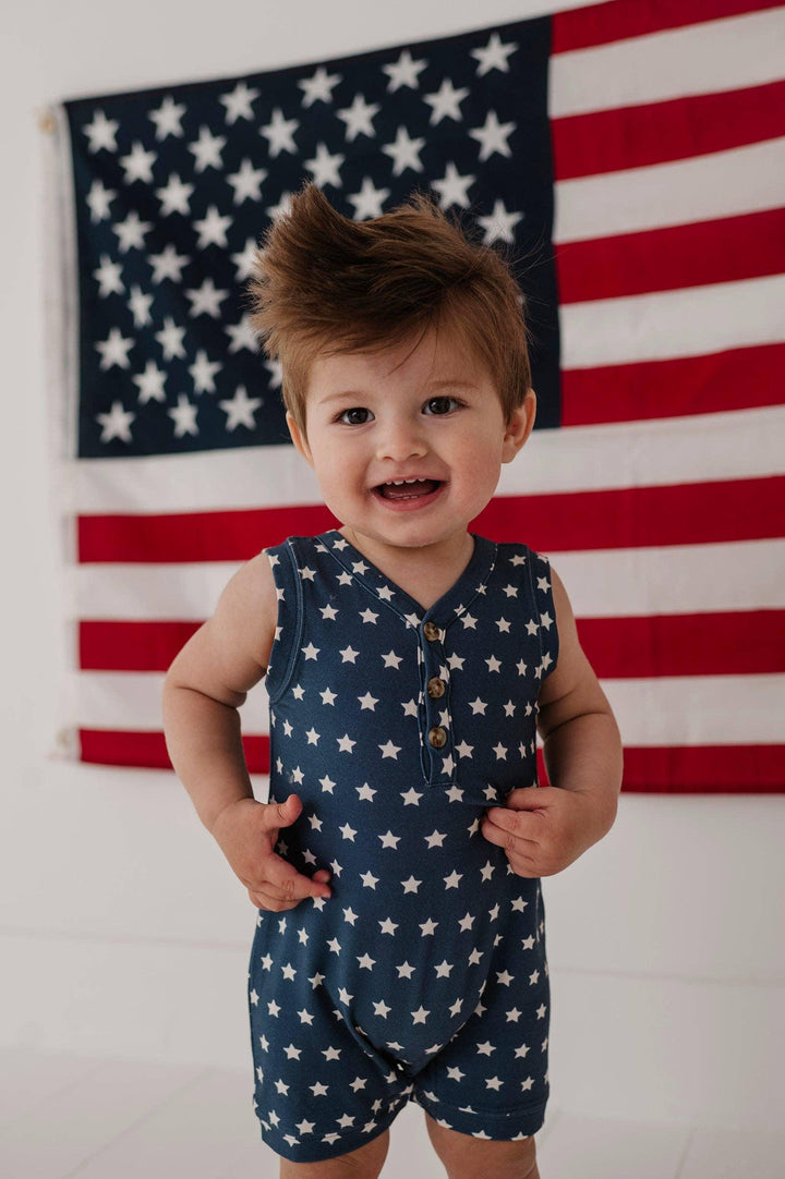 Baby in star-print romper smiling in front of American flag, perfect for 4th of July or patriotic adventures