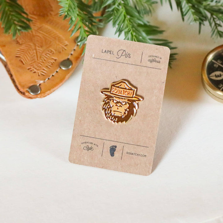 Squatchy lapel pin with forest ranger hat, designed in California, displayed with leather goods and pine branches
