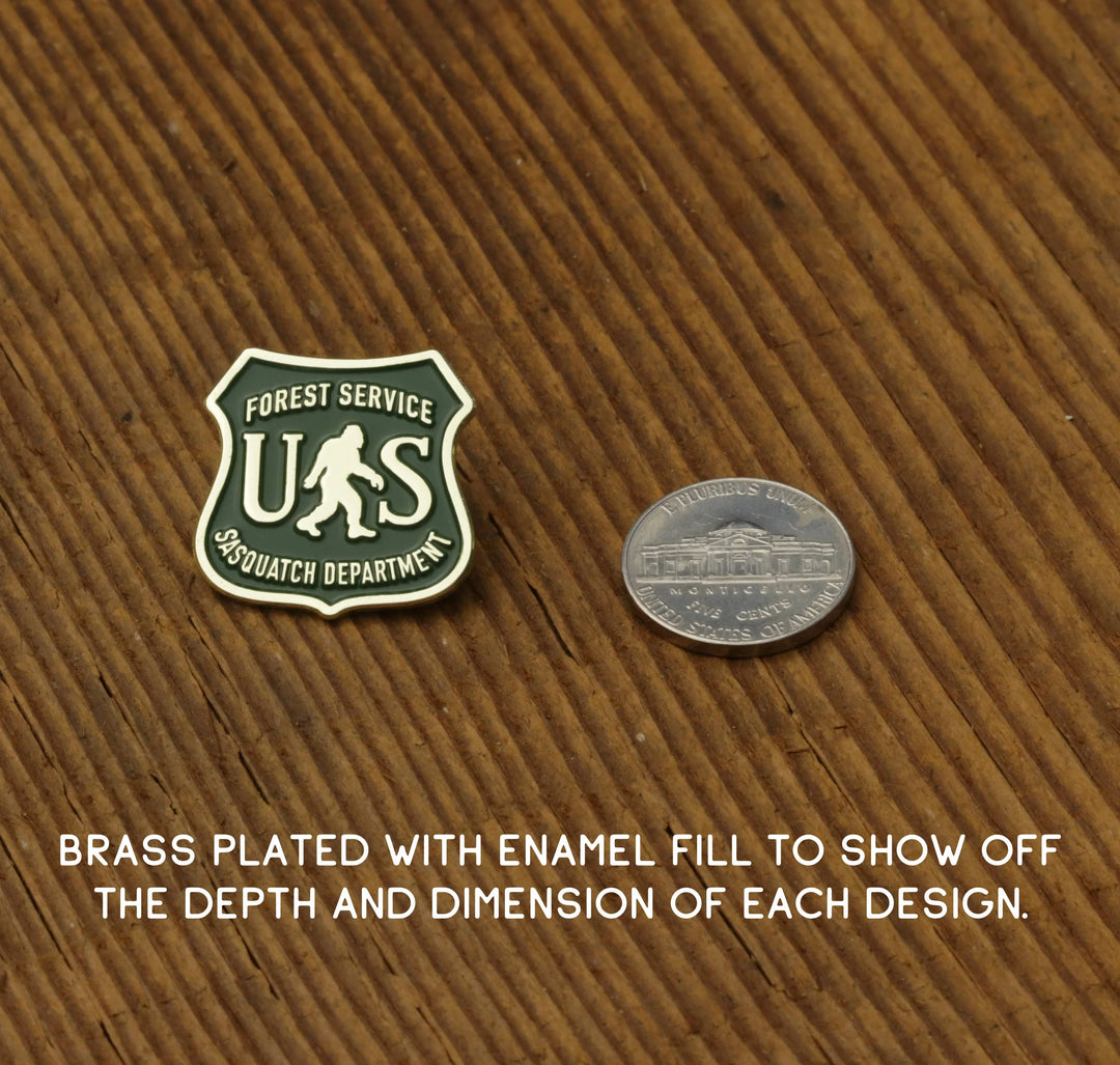 Forest Service Sasquatch Department enamel pin with brass plating, shown next to nickel for scale on wood