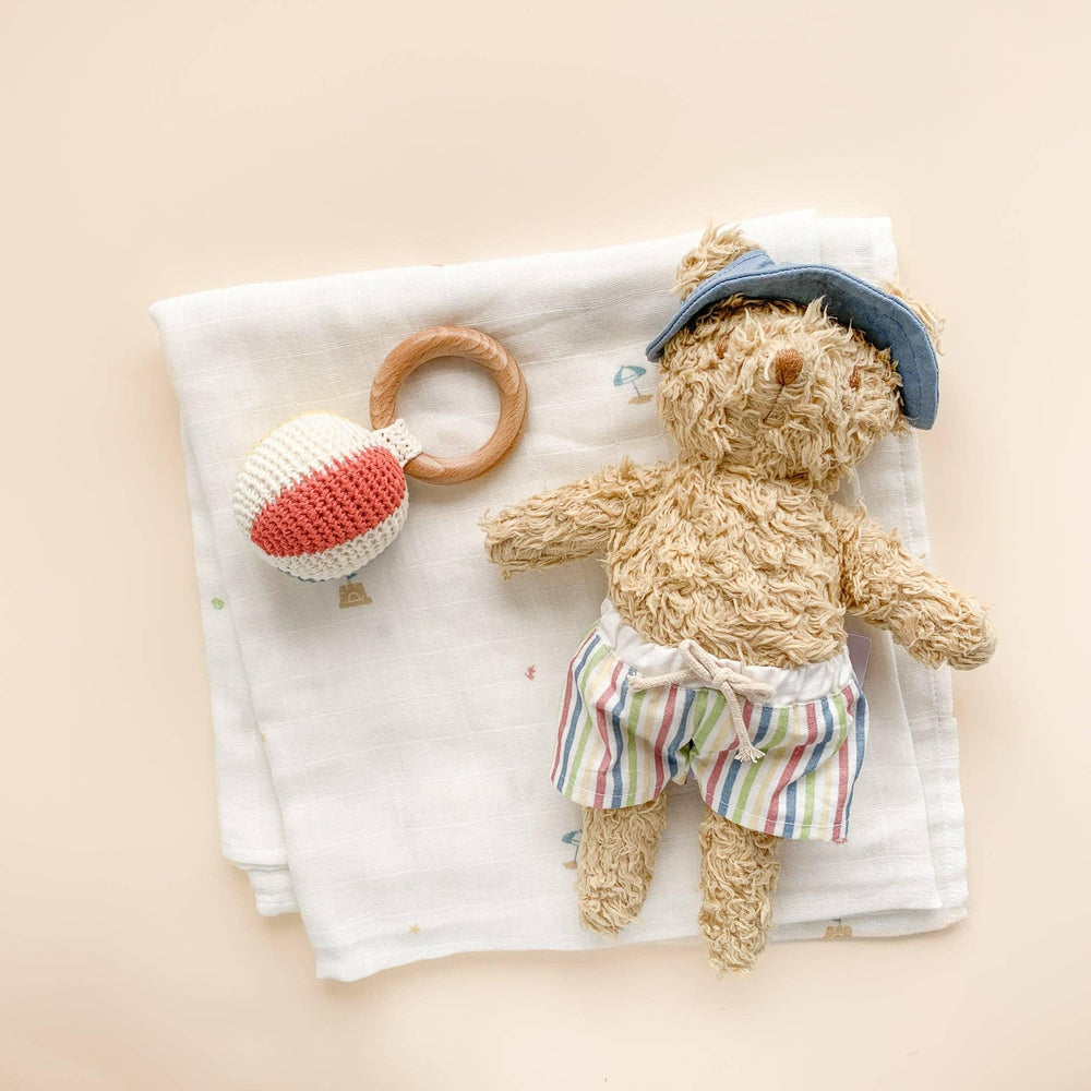 Cozy teddy bear in striped shorts and blue hat with knit hot air balloon rattle on muslin blanket