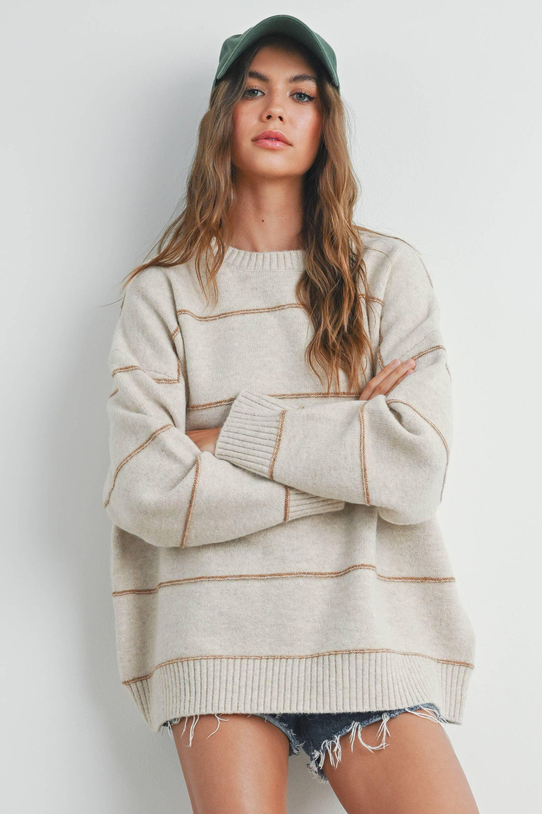 Woman in cozy cream sweater with tan stripes, green cap, and denim shorts