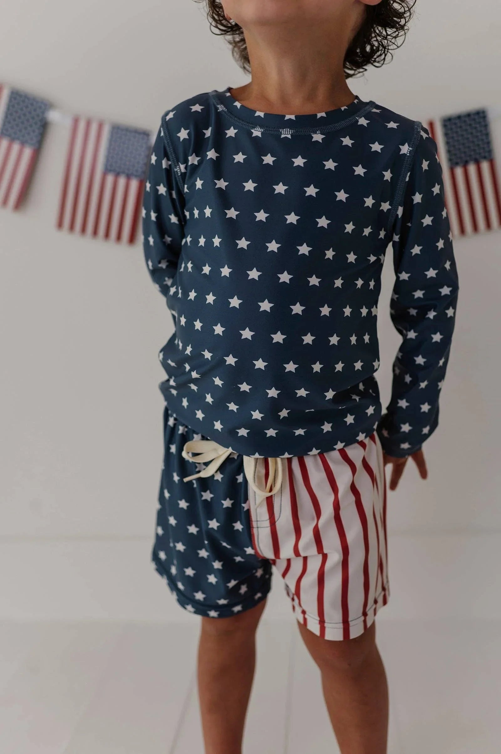 Kid in navy star rash guard and red stripe swim trunks, patriotic beachwear for coastal adventures