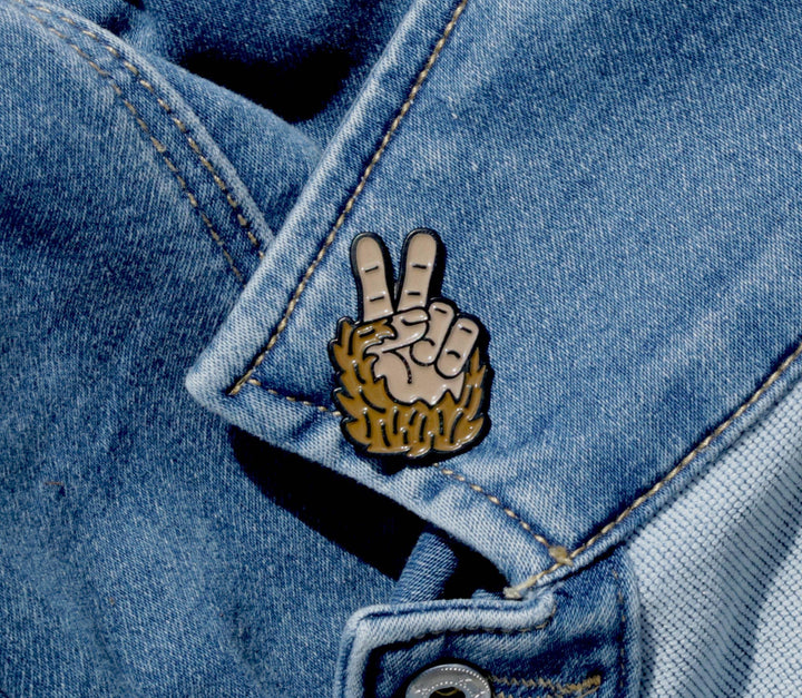 Squatchy enamel pin of a Bigfoot hand making a peace sign on a blue denim jacket.