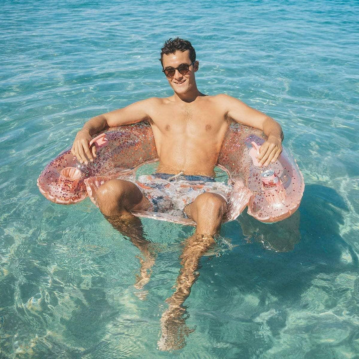Man relaxing on glittery pink pool float in clear blue water, perfect for beach days and coastal adventures.