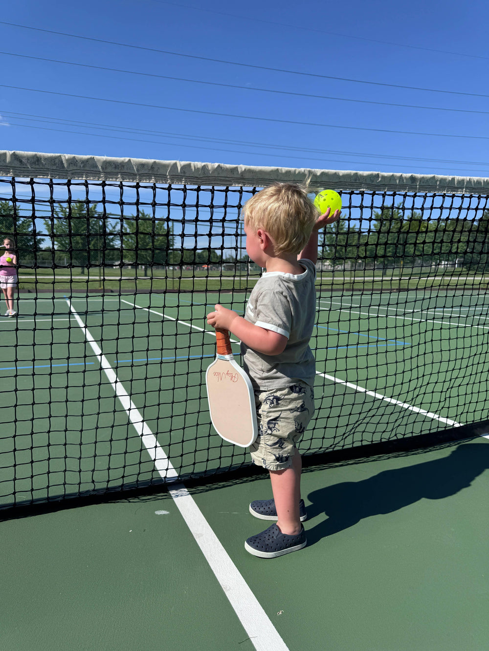 Toddler holding pickleball paddle and ball on outdoor court, ready to play under sunny sky