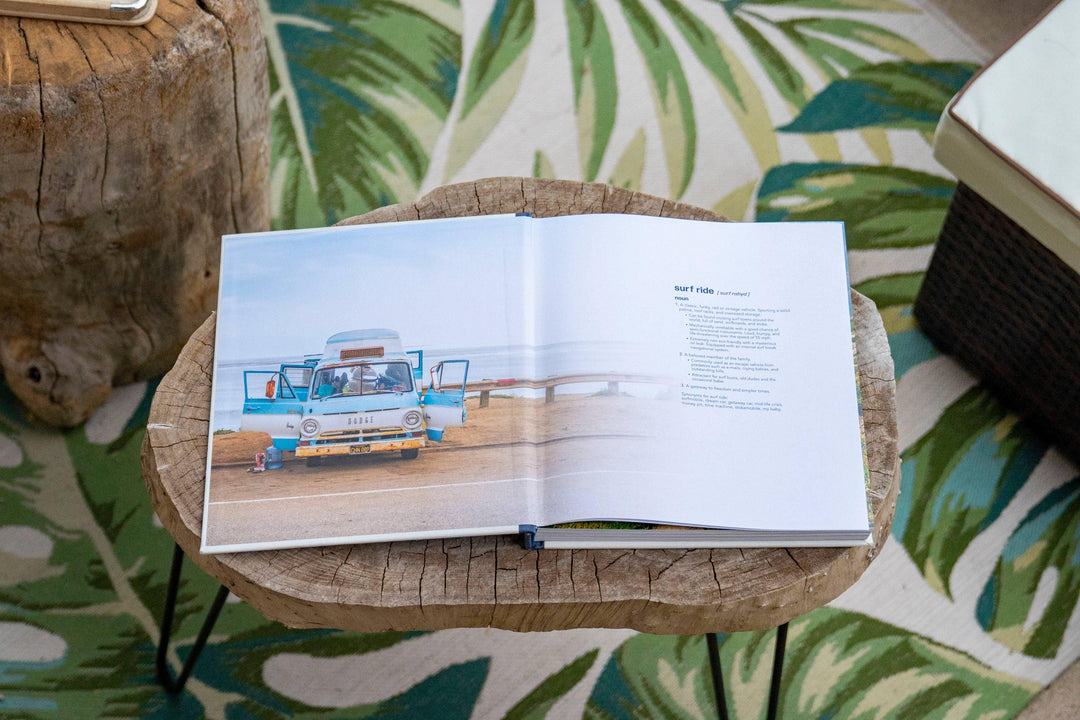 Open book on wooden table showing vintage surf van, coastal lifestyle guide with palm leaf rug nearby