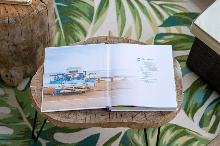 Open book on wooden table showing vintage surf van, coastal lifestyle guide with palm leaf rug nearby