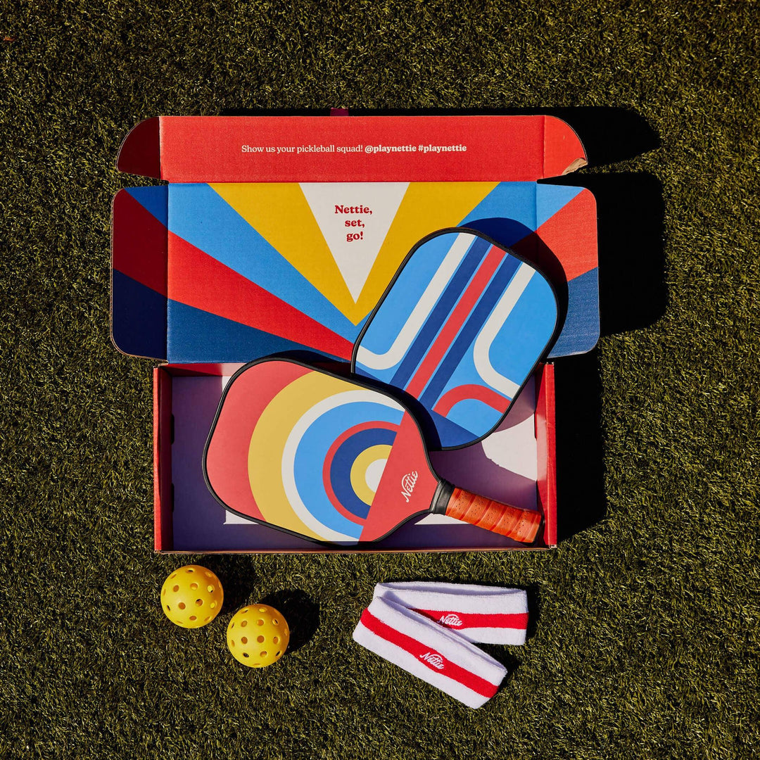 Colorful Nettie pickleball paddles, balls, and wristbands in open box on grass, ready for outdoor play.