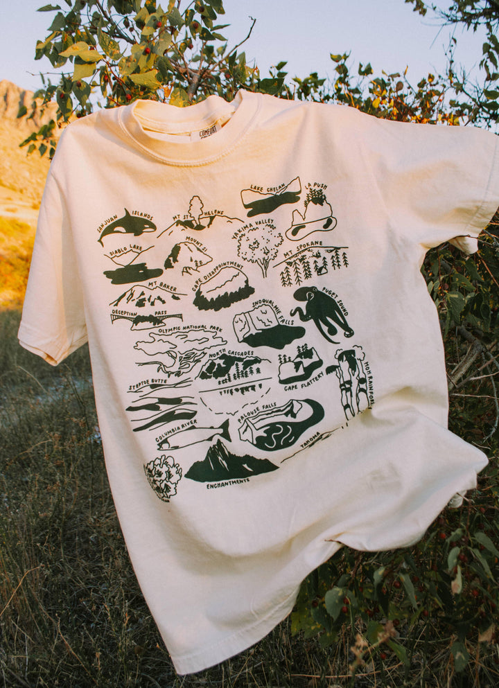 Cream t-shirt with PNW landmarks map graphic, laid outdoors in grass under tree