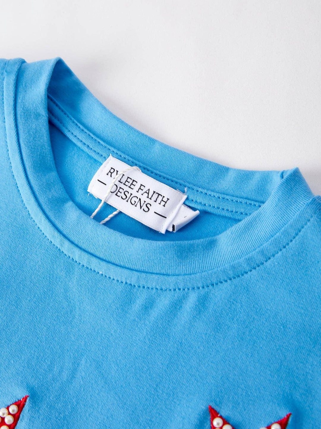 Bright blue t-shirt with red star appliqués and pearl details, featuring Rylee Faith Designs tag for coastal style