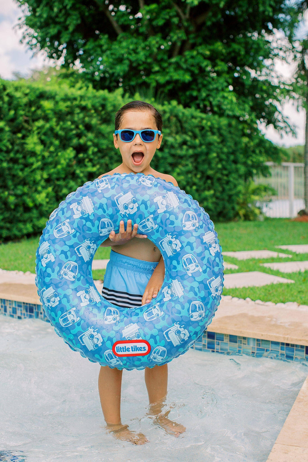 Boy in pool with blue car-patterned swim ring, wearing sunglasses and swim trunks