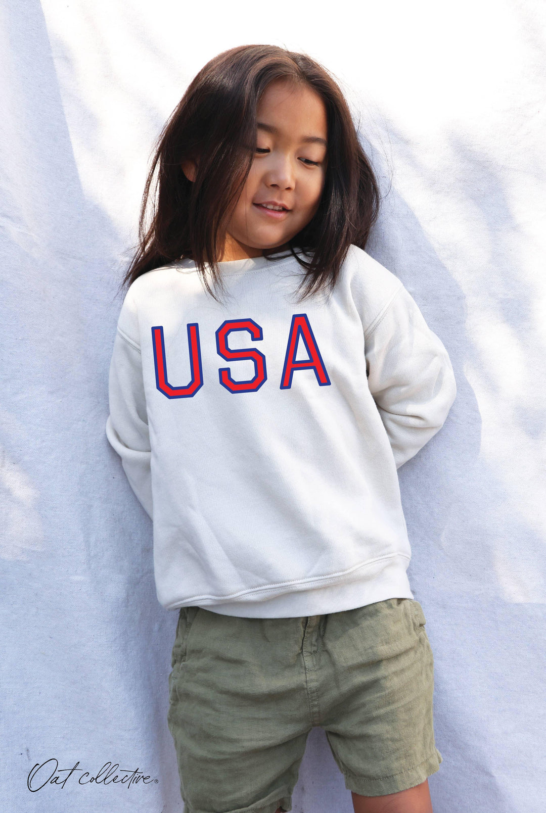 Child wearing white USA graphic sweatshirt and olive shorts, coastal outdoor style
