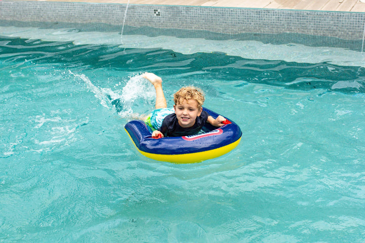 Boy riding blue and yellow pool float in backyard swimming pool, splashing water