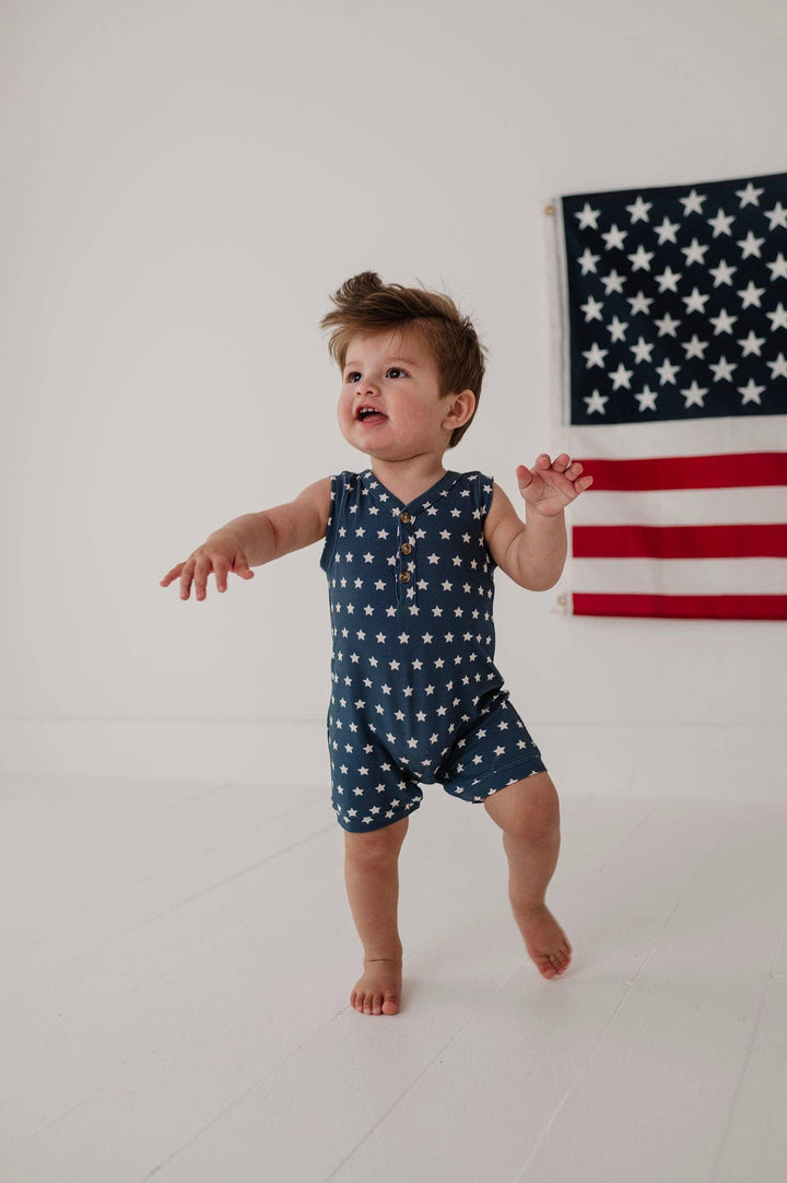 Baby in navy star print romper, standing barefoot near American flag, playful summer outfit