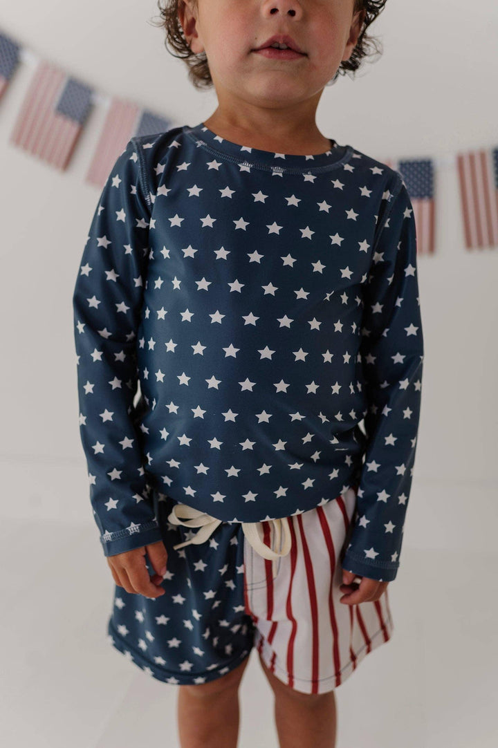 Toddler in navy star print long sleeve rash guard and red white striped swim trunks