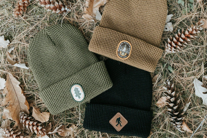 Three waffle knit beanies in olive, tan, and black with Pacific Northwest patches, nestled in pine needles and cones.