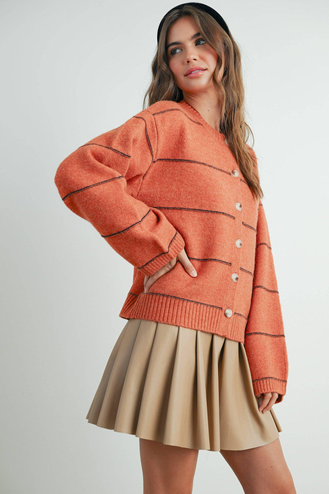 Woman in cozy orange cardigan with contrast stitching, paired with beige pleated skirt and black headband