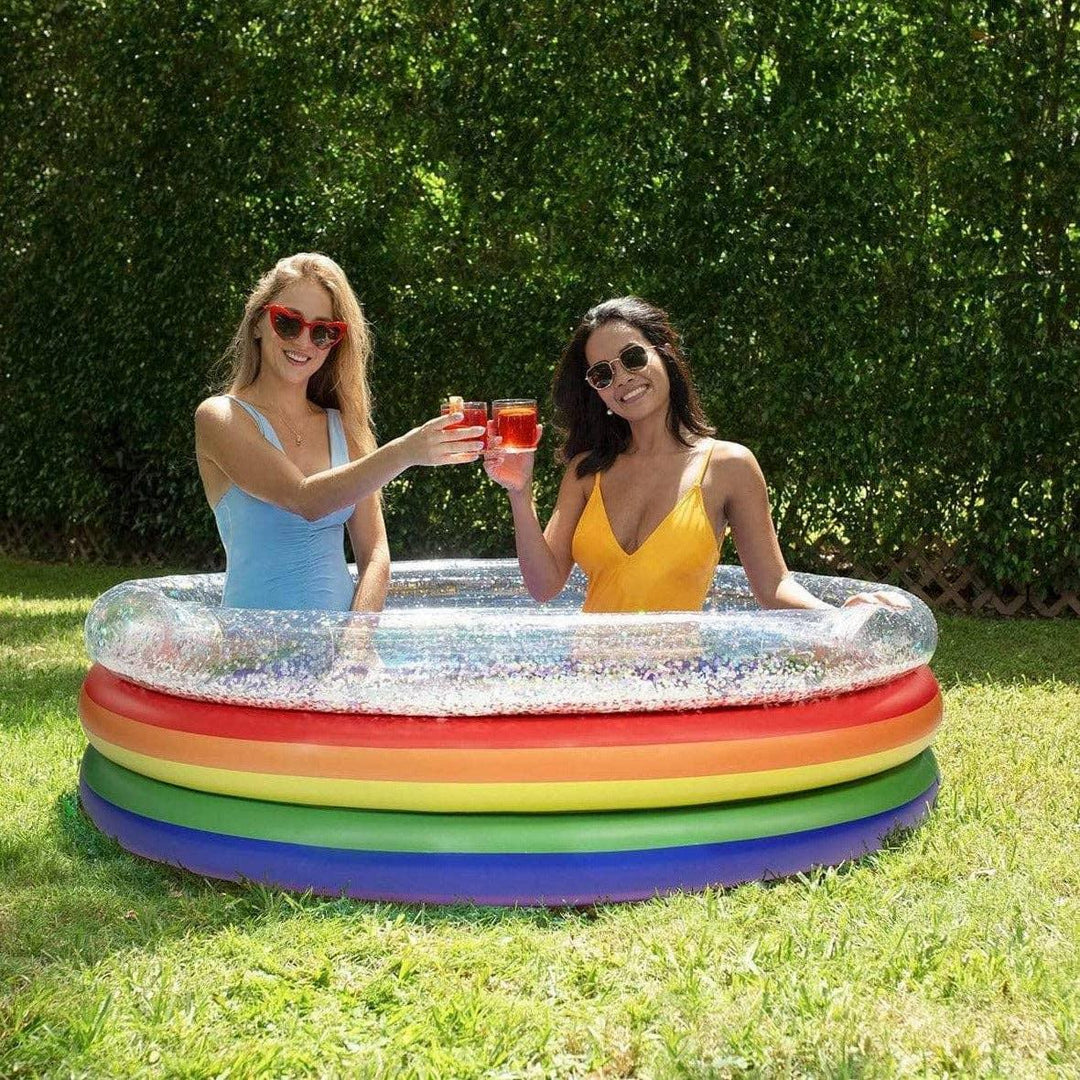 Two women toasting in a rainbow inflatable pool on a sunny lawn, perfect for backyard fun and summer vibes.