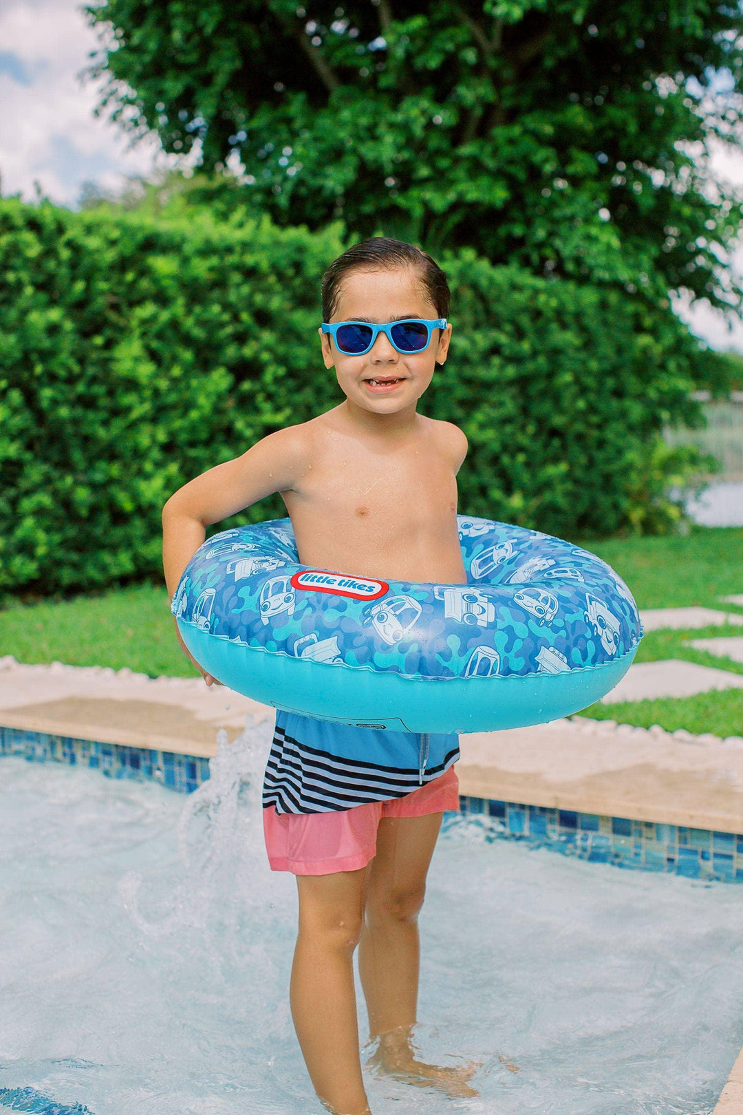 Child wearing sunglasses holding blue inflatable swim ring by pool