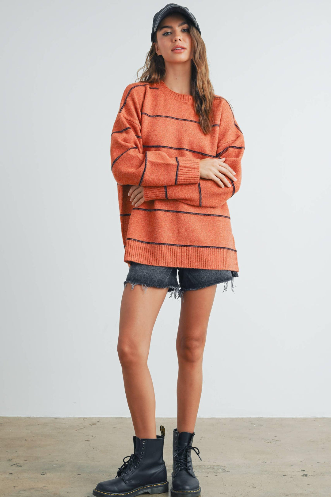 Woman in orange striped sweater, denim shorts, black boots, and cap, standing with arms crossed