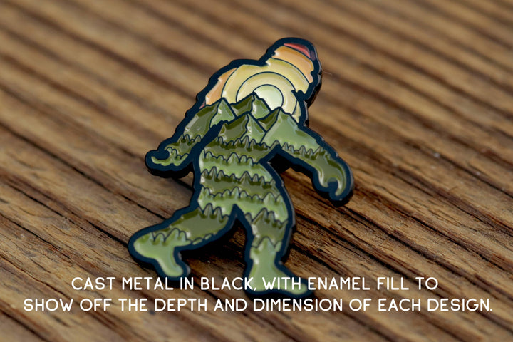 Cast metal enamel pin featuring a mountain-themed Bigfoot design on wooden surface