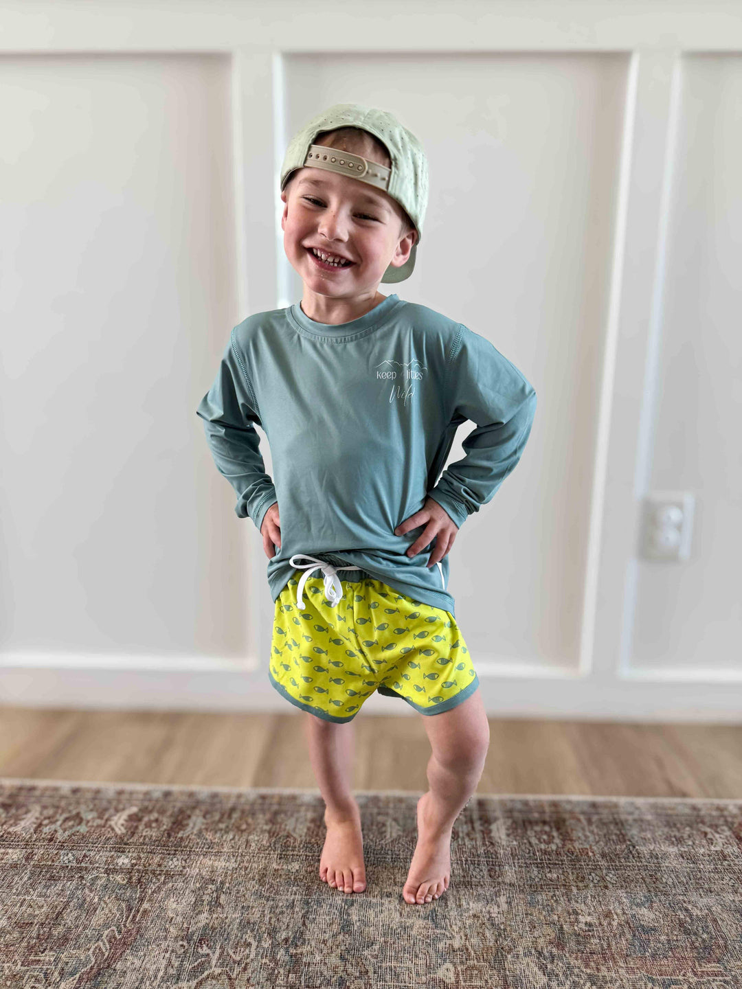 Smiling boy in teal long-sleeve shirt and fish-print swim shorts, ready for beach days and outdoor adventures.