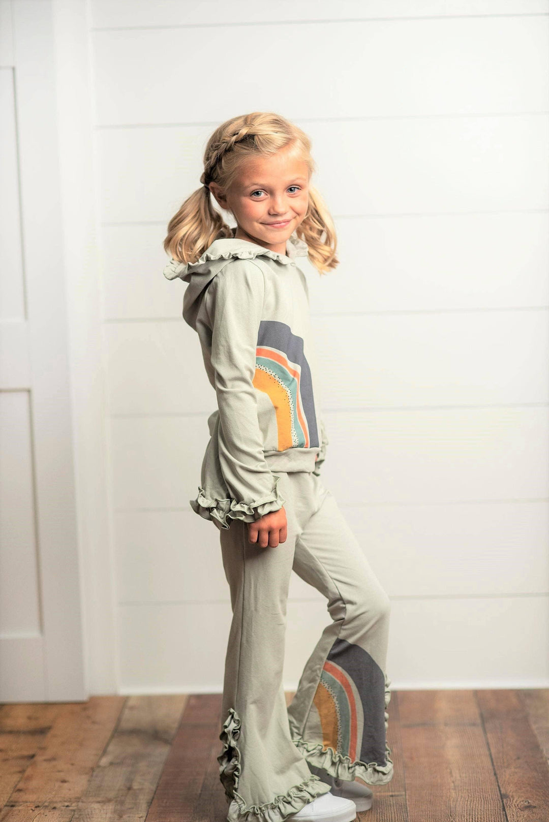 Girl wearing Oopsie Daisy gray sage rainbow hooded sweatsuit lounge set with ruffle trim.