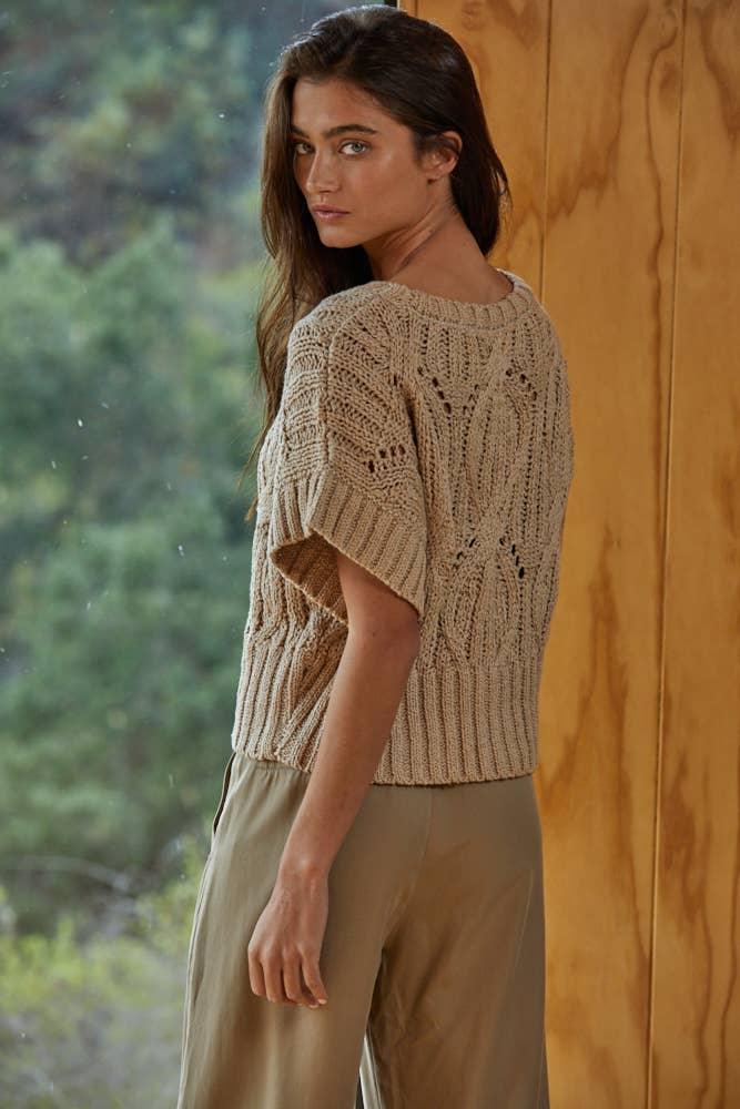 Woman wearing beige knit short-sleeve sweater with textured pattern, standing by wooden wall and window