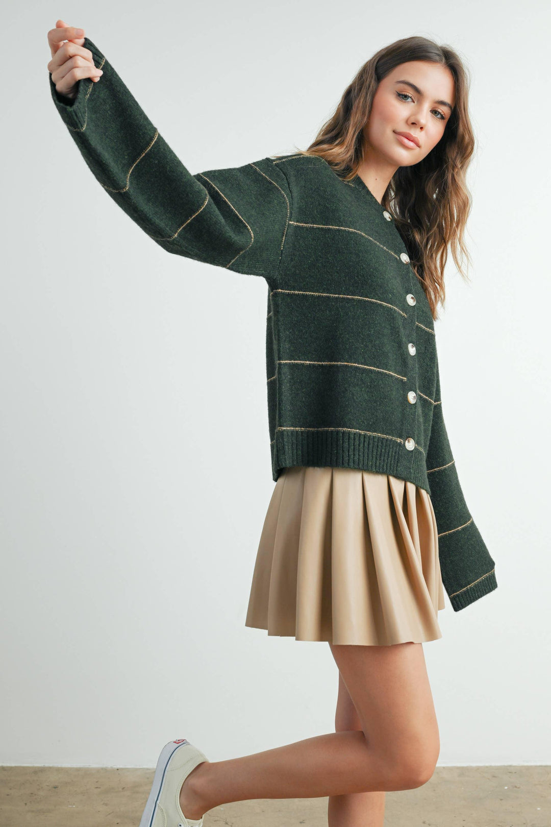 Woman in forest green cardigan with gold stripes, pleated skirt, and casual sneakers for coastal style