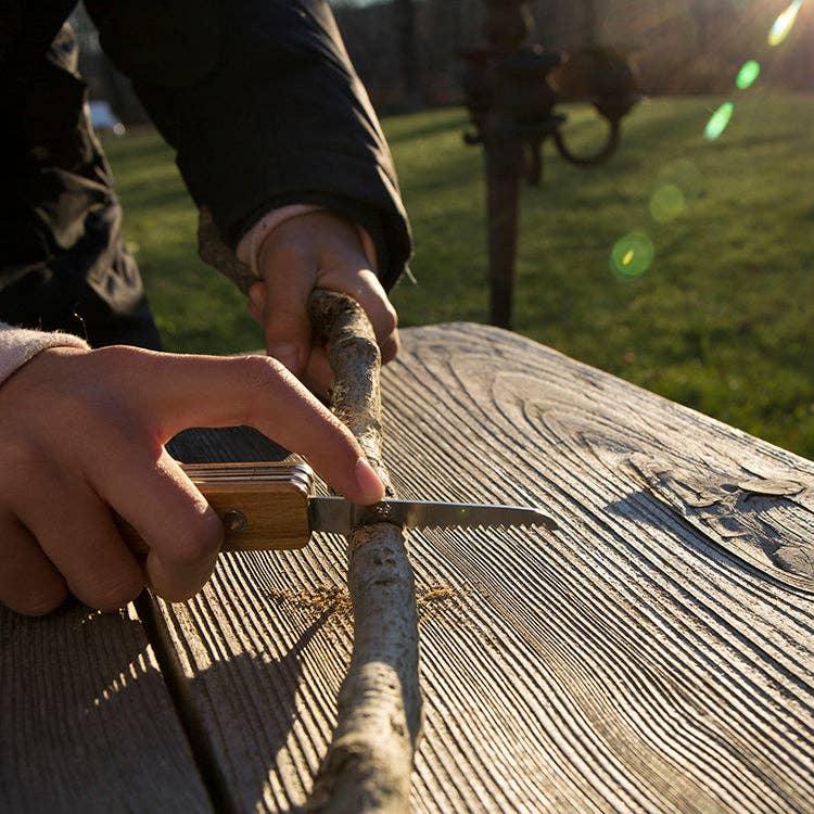 Hands using pocket saw to cut branch on wooden table outdoors, perfect for PNW adventures and outdoor gear.