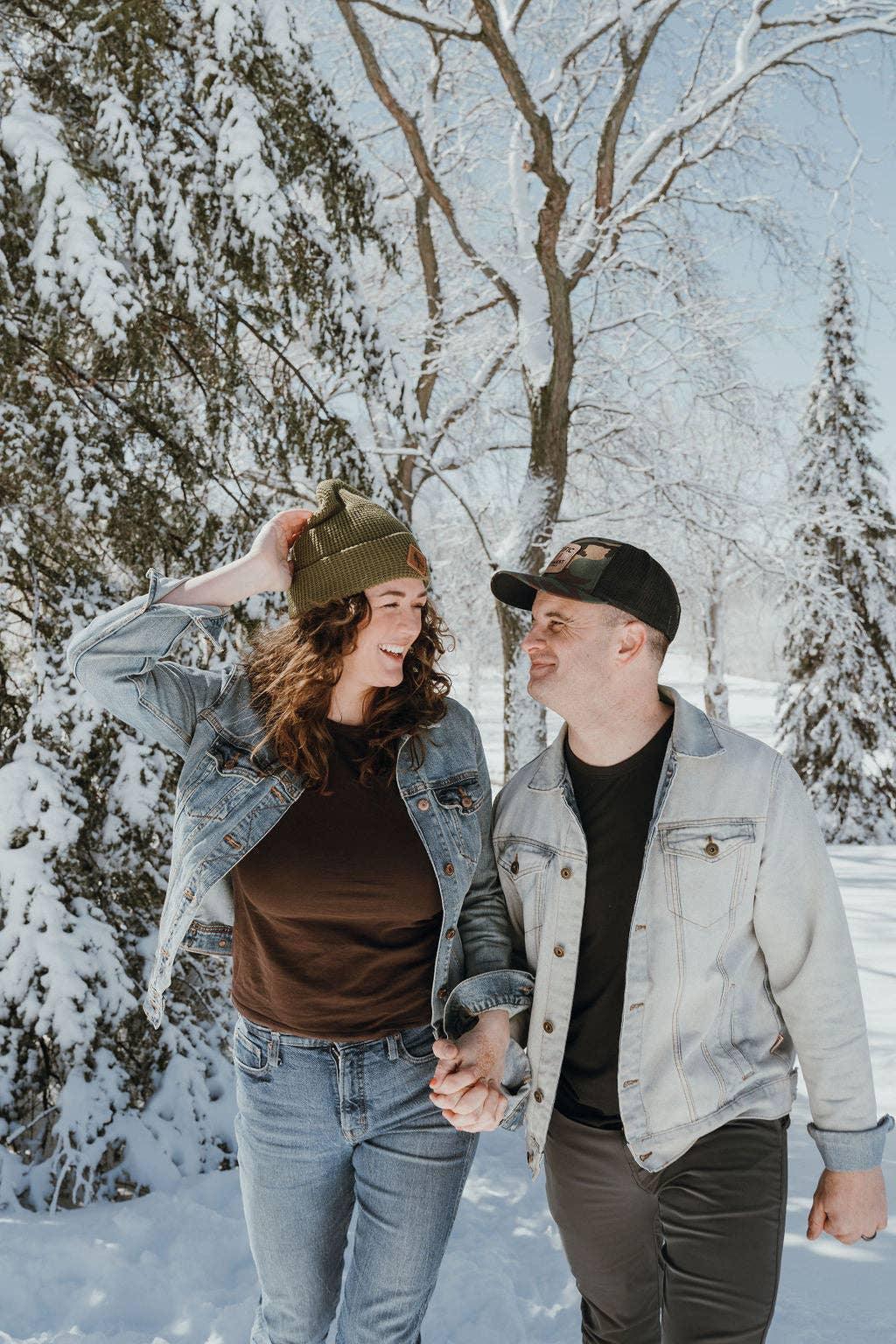 Couple in denim jackets holding hands in snowy forest, wearing beanies and outdoor gear